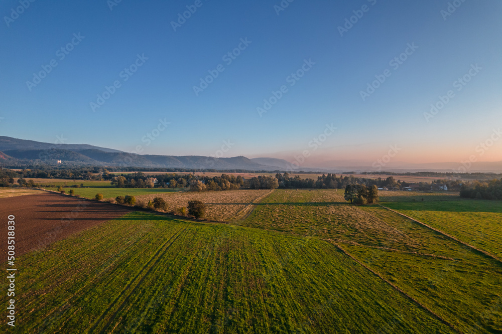 Green fields surrounded by the trees during sunrise. Blue sky and pastel colors. Mountains in the background.