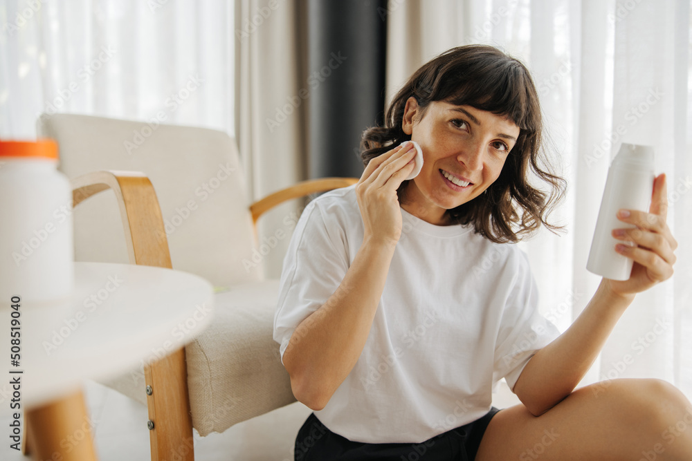 Happy young caucasian woman takes care of face skin, applies cleansing lotion after shower. Brunette is looking at camera and is wearing white t-shirt. Happy young caucasian woman takes care of face skin, applies cleansing lotion after shower. Brunette is looking at camera and is wearing white t-shirt.