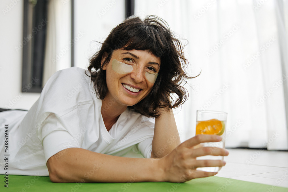 Joyful young caucasian woman avoiding dehydration by drinking water and using patches lying on mat. Brunette in white t-shirt looks at camera smiling. Skin care and hydration concept Joyful young caucasian woman avoiding dehydration by drinking water and using patches lying on mat. Brunette in white t-shirt looks at camera smiling. Skin care and hydration concept