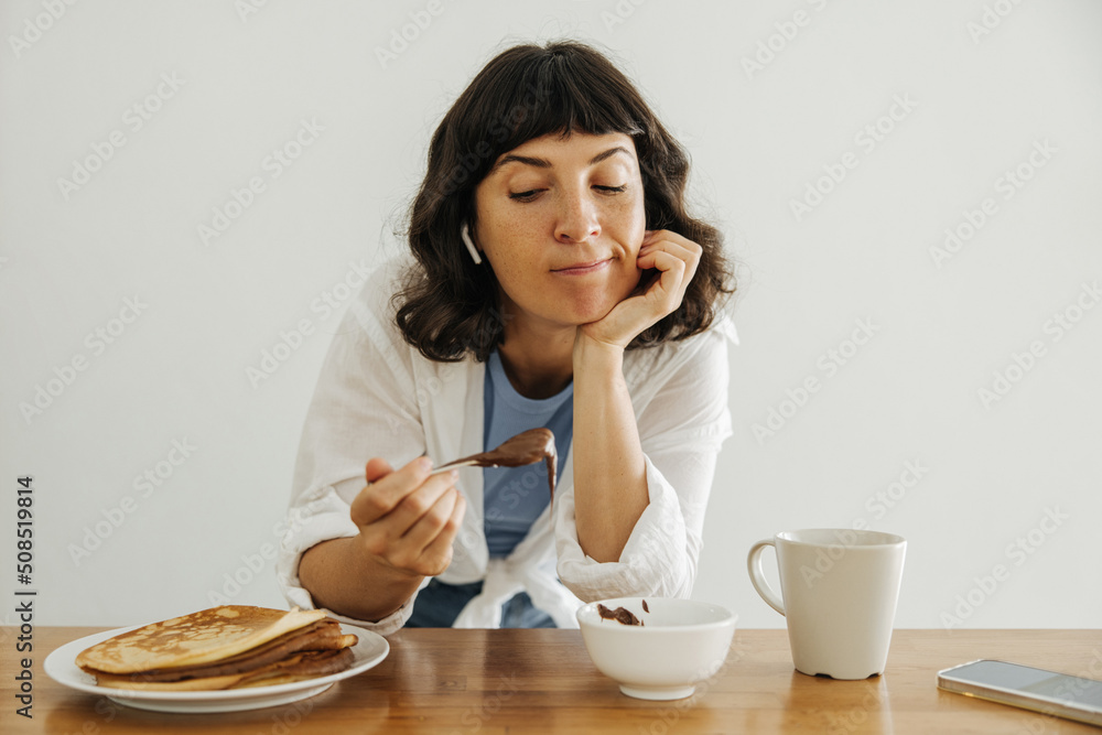 Unsure young caucasian woman relaxing at table enjoying delicious pancakes with chocolate. Brunette woman wears top, shirt and jeans at home. Breakfast concept Unsure young caucasian woman relaxing at table enjoying delicious pancakes with chocolate. Brunette woman wears top, shirt and jeans at home. Breakfast concept