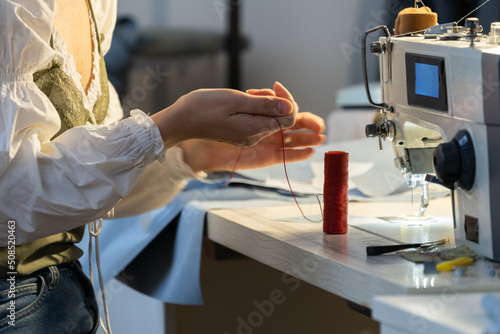 Young dressmaker woman sew clothes on sewing machine holding a spool of thread. Professional seamstress work in workshop create new product. Self employed atelier owner in studio. Small business photo