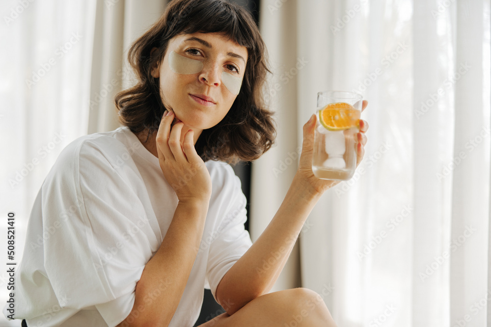 Attractive young caucasian girl in white t-shirt takes care of her facial skin and holds glass of water near window. Brunette conducts morning rituals at home. Health care concept Attractive young caucasian girl in white t-shirt takes care of her facial skin and holds glass of water near window. Brunette conducts morning rituals at home. Health care concept