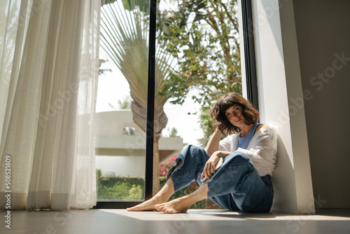 Pretty young caucasian woman is sitting by window and looking at camera holding mobile phone. Brunette with short haircut wears shirt and jeans. Lifestyle concept