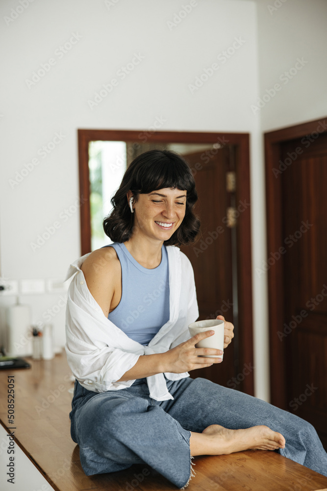 Pretty young caucasian woman with cup of coffee listens to music on headphones sitting in living room morning. Brunette with closed eyes wears casual clothes. Mood, lifestyle, concept. Pretty young caucasian woman with cup of coffee listens to music on headphones sitting in living room morning. Brunette with closed eyes wears casual clothes. Mood, lifestyle, concept.
