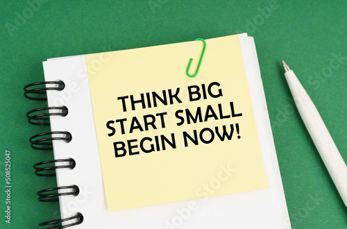 On a green surface, a pen, a notepad with stickers and the inscription - Think Big Start Small Begin Now