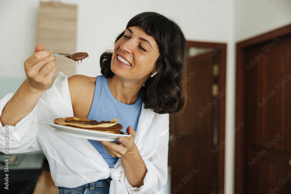 Positive young caucasian girl is enjoying delicious breakfast of pancakes with chocolate. Brunette with short haircut wears casual clothes at home. Good morning concept Positive young caucasian girl is enjoying delicious breakfast of pancakes with chocolate. Brunette with short haircut wears casual clothes at home. Good morning concept
