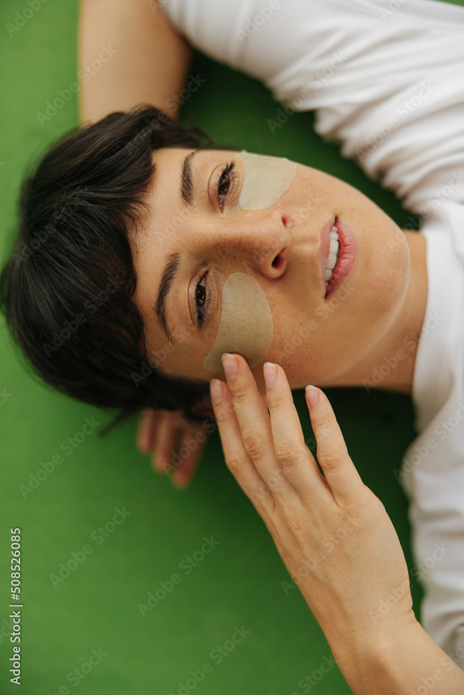Pleased young caucasian woman applying green hydrogel pads under eyes for skin treatment lying on mat. Brunette looks in front of her and touches face. Cosmetic natural trend Pleased young caucasian woman applying green hydrogel pads under eyes for skin treatment lying on mat. Brunette looks in front of her and touches face. Cosmetic natural trend