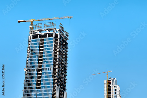 Yellow tower crane on the construction of a modern skyscraper with windows and a white skyscraper under construction with a crane behind against a clear blue sky in bright sunlight