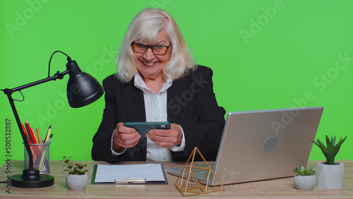 Fotografie Worried funny senior businesswoman sits at workplace avoid working and enthusiastically playing racing or shooter video games at office desk table