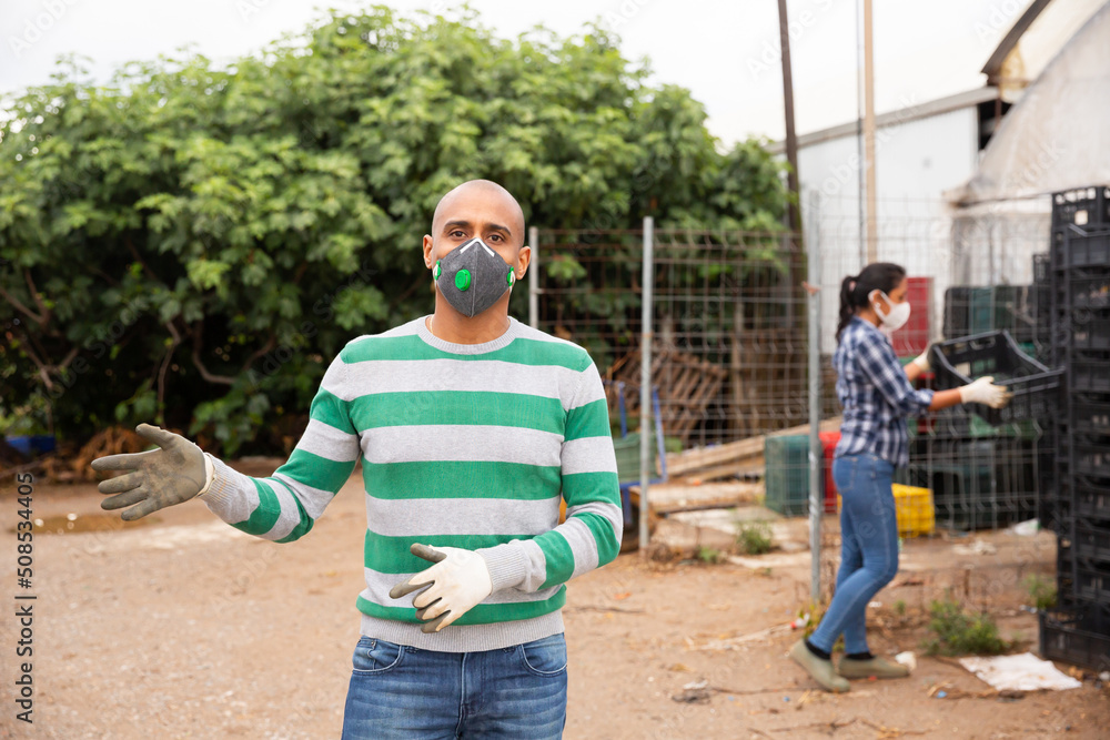 Naklejka premium Confident hispanic man farm worker in protective face mask standing outside near greenhouse buildings
