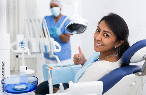 Happy latin american woman sitting in dental chair after teeth cure in modern clinic, gesturing thumbs up