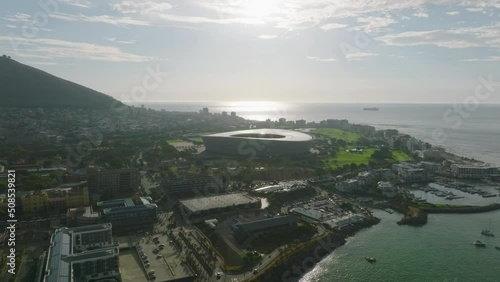 Aerial view of sea coast and buildings on waterfront. Modern circular structure of football stadium against setting sun. Cape Town, South Africa photo