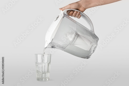 Woman pouring purified water into glass from filter jug on grey background