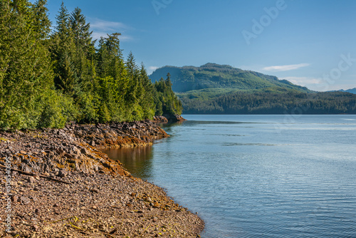 Ketchikan, Alaska, USA - July 17, 2011: Landscape, dense green forests above brown rocky shoreline along Tongass Narrows under blue sky. Mountains on horizon and blue ocean water up front. photo