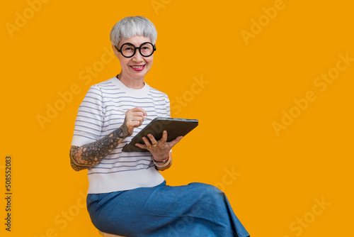 smiling retired old senior asian female with tattoo relax on comfy stool use modern digital touchpad tablet device read ebook from screen scroll social media web pages isolate yellow background
