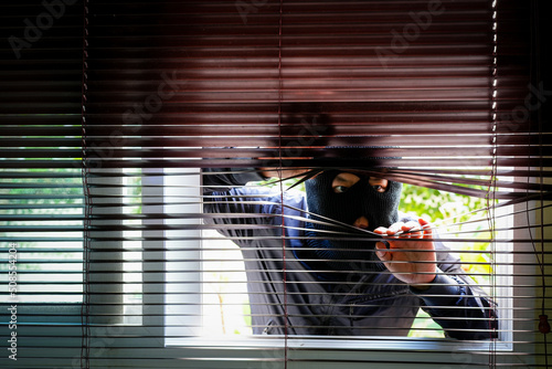 Unrecognizable Man Wearing Balaclava Face Mask Looking Through Venetian Blind. Keep your home secure during your summer holiday Concept.