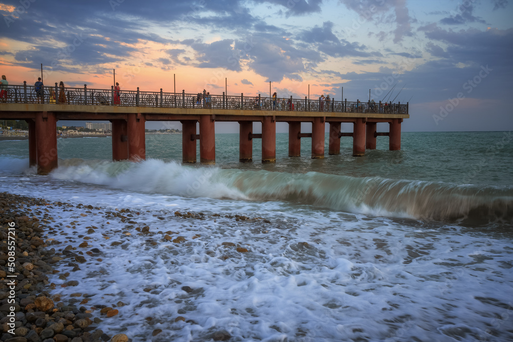 Evening walk along the old high pier over the sea in Adler (Russia) in summer. Waves crash against shore, forming foam