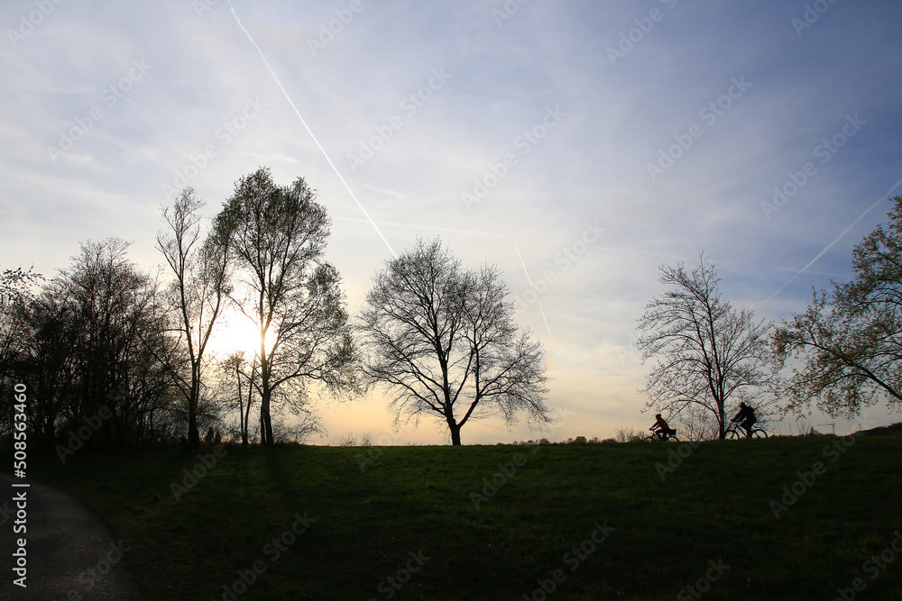 Regensburg, Germany: silhouette of people riding the bike on a rural road at sunset along Danube river in Regensburg, Germany, Europe. Regensburg, Germany: silhouette of people riding the bike on a rural road at sunset along Danube river in Regensburg, Germany, Europe.