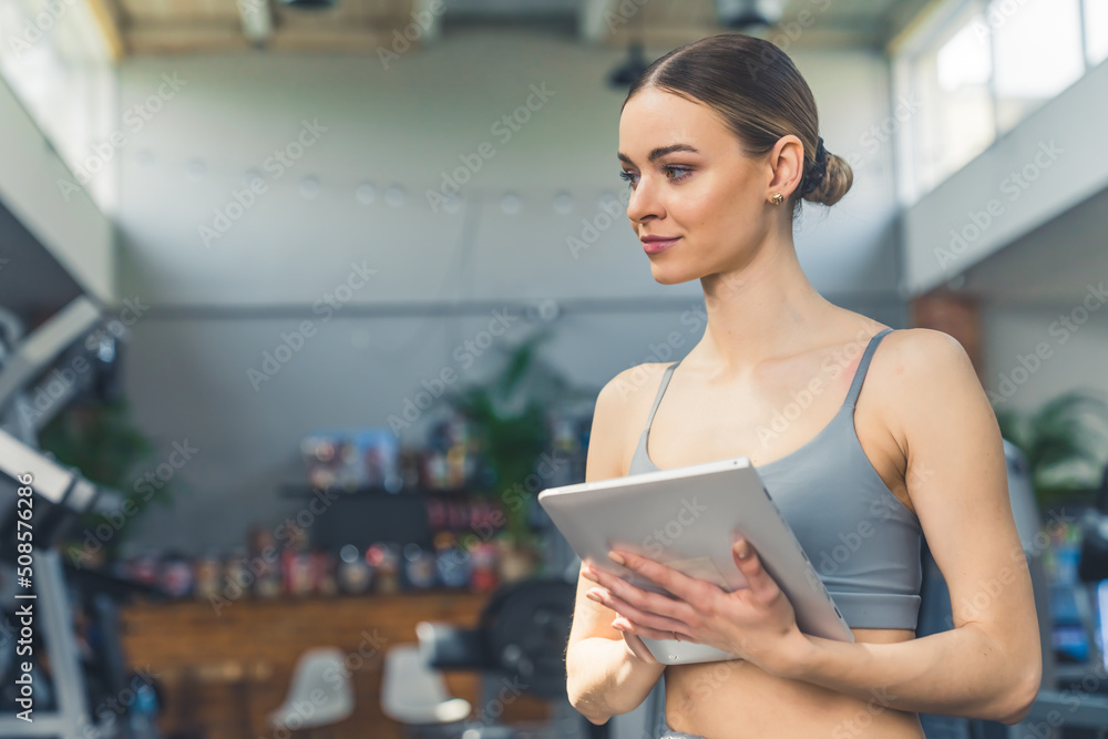 Indoor portrait of a young white millennial woman with slicked back hair and gray sports bra holding a tablet, creating workout program for her clients. High quality photo Indoor portrait of a young white millennial woman with slicked back hair and gray sports bra holding a tablet, creating workout program for her clients. High quality photo