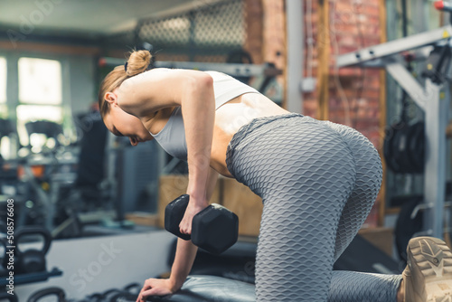 Time for muscle building. Young caucasian girl leaning on a black bench and doing triceps exercises with the use of a black dumbbell. High quality photo
