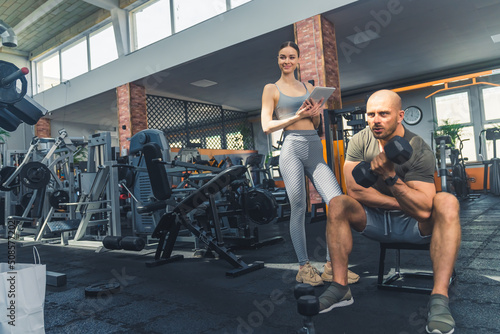 Female caucasian personal trainer holding a tablet and acknowledging the progress of her fellow bodybuilding colleague. Gym interior. High quality photo