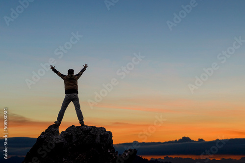 Success concept.Silhouette male hiker with his hands raised enjoy sunny day on top rock mountain sunset background.