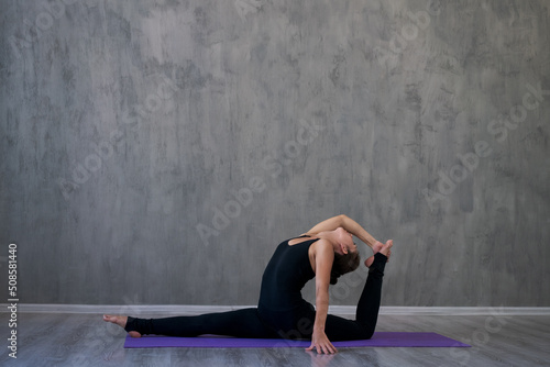 Attractive young woman doing yoga stretching yoga online at home against gray wall in black sportswear. Self-isolation is useful entertainment and education on Internet. The concept healthy lifestyle.