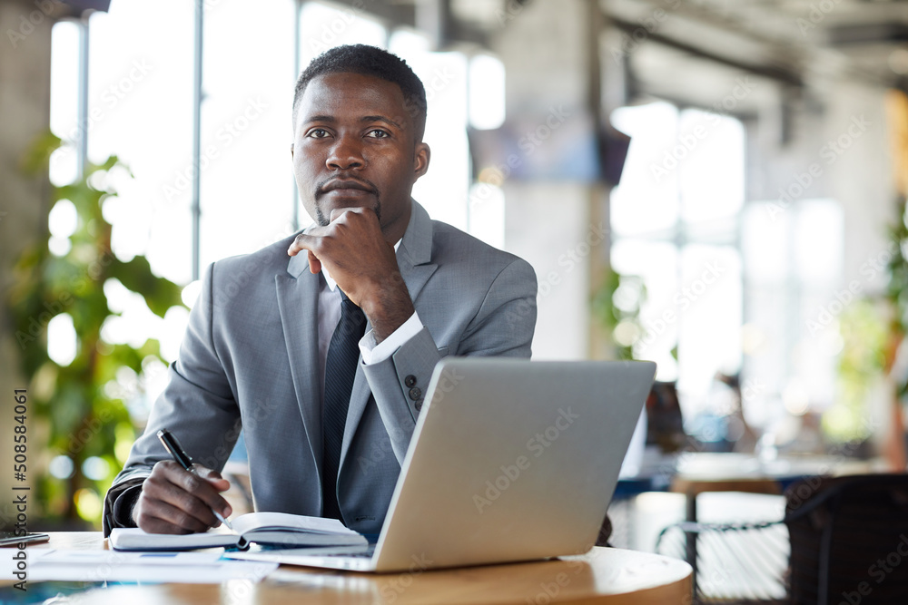 Serious handsome young black businessman in stylish suit sitting at table with diary and laptop and looking at camera in restaurant Serious handsome young black businessman in stylish suit sitting at table with diary and laptop and looking at camera in restaurant