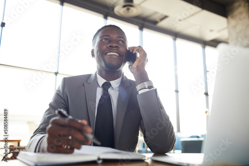 Cheerful confident young African-American businessman in formal suit sitting at table in restaurant and discussing project to colleague on mobile phone