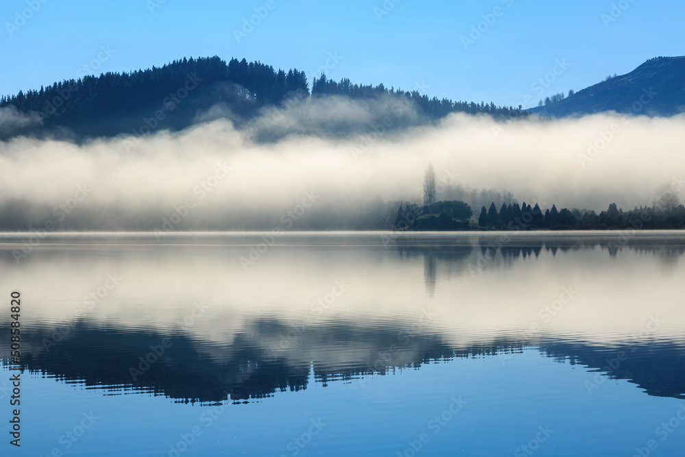 Fototapeta premium A blanket of mist reflected in the calm waters of a lake. Photographed at Lake Tutira, New Zealand