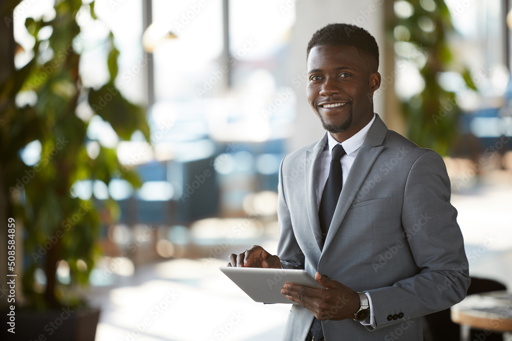 Positive purposeful young black business manager with beard standing in office lobby and using digital tablet while looking at camera Positive purposeful young black business manager with beard standing in office lobby and using digital tablet while looking at camera