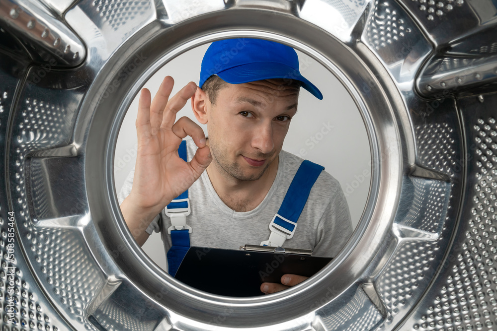 Male repairman in uniform with a tablet in his hands shows the OK sign, looks into the drum of the washing machine, photo from the inside. Male repairman in uniform with a tablet in his hands shows the OK sign, looks into the drum of the washing machine, photo from the inside.