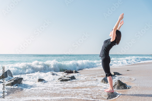 Side view of female stretching body and doing backbend while standing on rock in Tadasana. She is with raised arms on sea coast against blue sky during yoga session photo