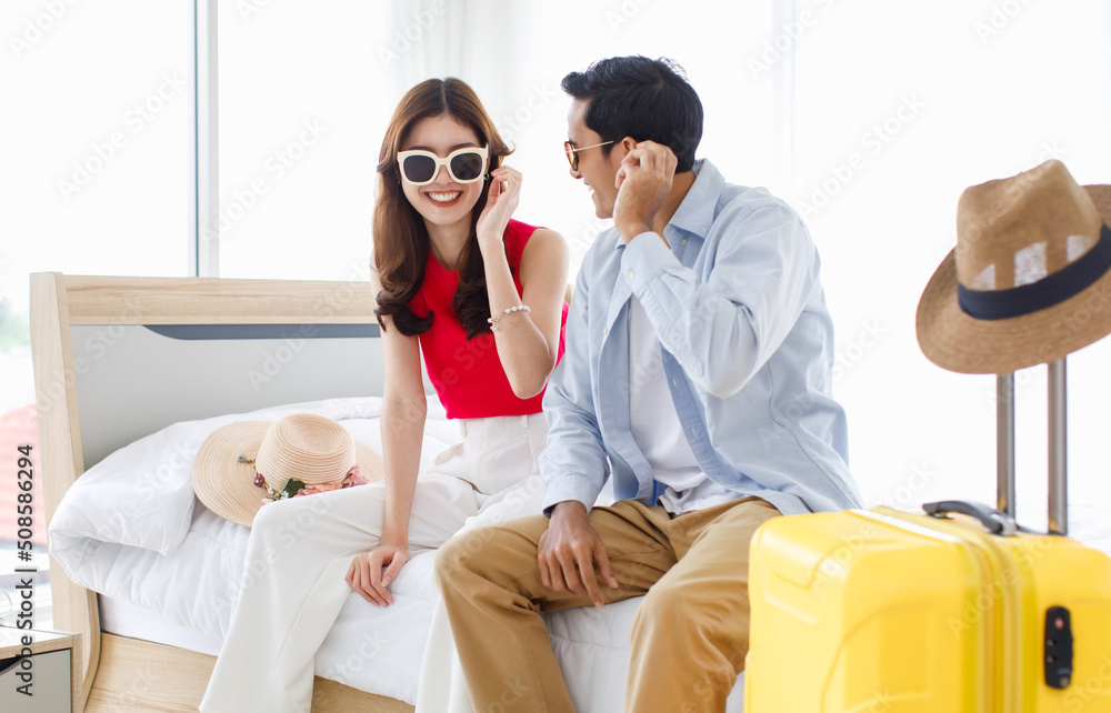 Obraz premium Asian young happy male female traveler lover couple husband wife in casual outfit with sunglasses and hat sitting smiling on bed in bedroom packing luggage ready for summer vacation trip together