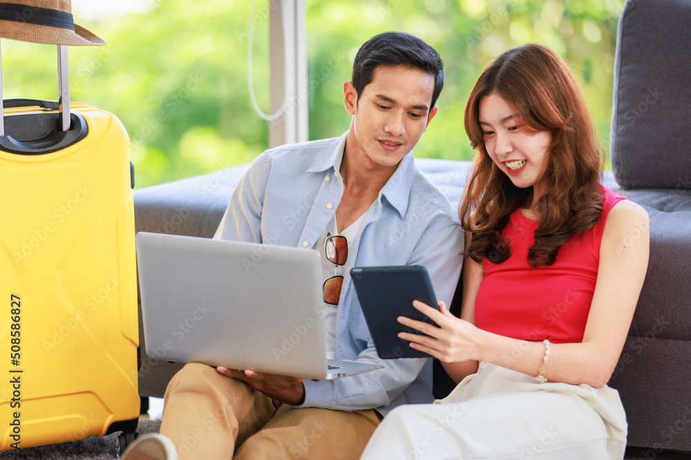 Asian young lovely boyfriend and girlfriend traveler couple sitting smiling on carpet in living room with trolley luggage while using laptop computer and tablet booking vacation summer trip online Asian young lovely boyfriend and girlfriend traveler couple sitting smiling on carpet in living room with trolley luggage while using laptop computer and tablet booking vacation summer trip online