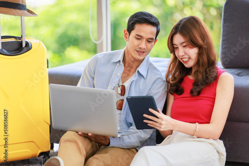 Asian young lovely boyfriend and girlfriend traveler couple sitting smiling on carpet in living room with trolley luggage while using laptop computer and tablet booking vacation summer trip online