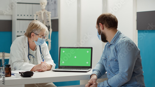 Doctor and patient looking at laptop with greenscreen at chekcup visit in cabinet. Physician and man using blank copyspace mockup with isolated chromakey template during covid 19 pandemic.