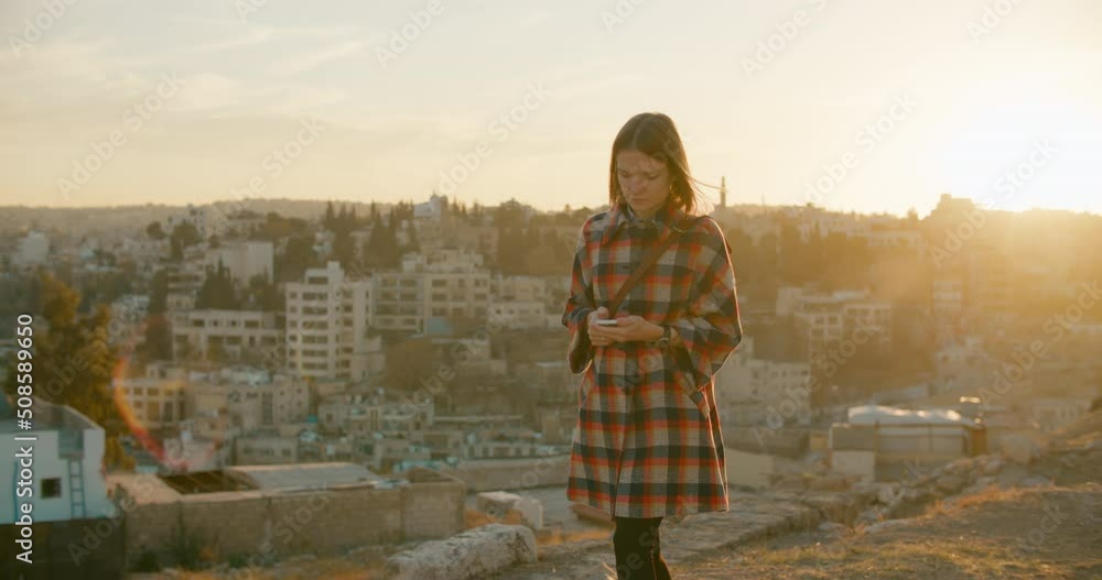 Young Lady uses Mobile Smartphone App in Amman, Jordan, with typical Middle East Cityscape. Female Tourist Woman texts message walking outdoors with Jordanian Skyline at sunset. 4K wide tracking shot Young Lady uses Mobile Smartphone App in Amman, Jordan, with typical Middle East Cityscape. Female Tourist Woman texts message walking outdoors with Jordanian Skyline at sunset. 4K wide tracking shot