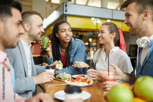 Group of cheerful young multi-ethnic students in casual clothing sitting at table on food court and having lunch together