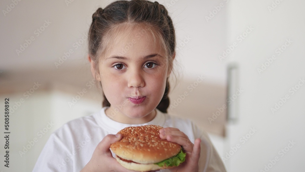 little girl eating a hamburger. unhealthy fast food proper nutrition concept. child greedily with pleasure bites a big burger in the kitchen at home. kid eats fast food lifestyle close-up little girl eating a hamburger. unhealthy fast food proper nutrition concept. child greedily with pleasure bites a big burger in the kitchen at home. kid eats fast food lifestyle close-up
