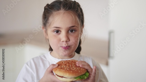 little girl eating a hamburger. unhealthy fast food proper nutrition concept. child greedily with pleasure bites a big burger in the kitchen at home. kid eats fast food lifestyle close-up