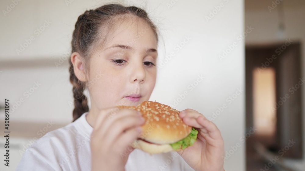 little girl eating a hamburger. unhealthy fast food proper nutrition concept meal. child greedily with pleasure bites a big burger in the kitchen at home. kid eats fast food close-up little girl eating a hamburger. unhealthy fast food proper nutrition concept meal. child greedily with pleasure bites a big burger in the kitchen at home. kid eats fast food close-up