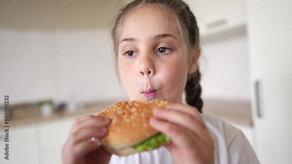 little kid girl eating a hamburger. unhealthy fast food meal proper nutrition concept. child greedily with pleasure bites a big burger lifestyle in the kitchen at home. kid eats fast food close-up little kid girl eating a hamburger. unhealthy fast food meal proper nutrition concept. child greedily with pleasure bites a big burger lifestyle in the kitchen at home. kid eats fast food close-up