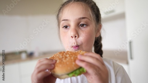 little kid girl eating a hamburger. unhealthy fast food meal proper nutrition concept. child greedily with pleasure bites a big burger lifestyle in the kitchen at home. kid eats fast food close-up