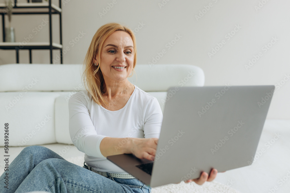 Mature woman using laptop on floor with a happy face standing and smiling with a confident smile showing teeth Mature woman using laptop on floor with a happy face standing and smiling with a confident smile showing teeth