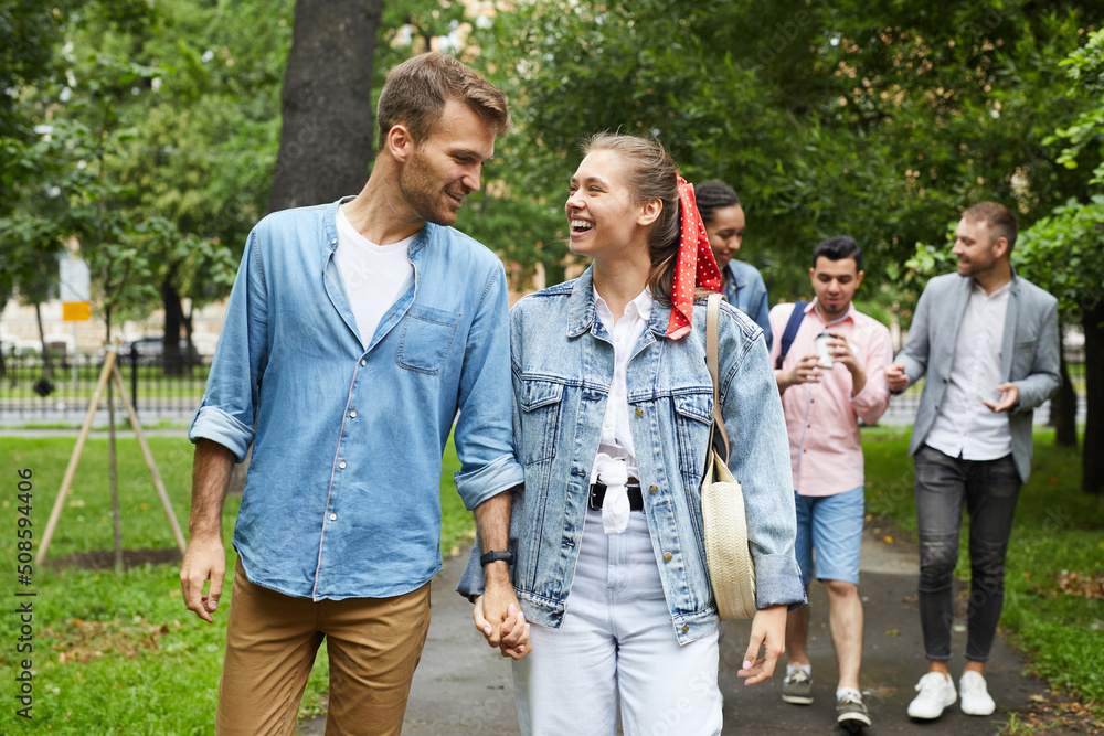 Happy beautiful young student couple in casual outfits holding hands and talking while strolling in park Happy beautiful young student couple in casual outfits holding hands and talking while strolling in park