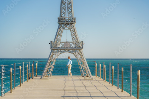 Large model of the Eiffel Tower on the beach. A woman walks along the pier towards the tower, wearing a blue jacket and white jeans.