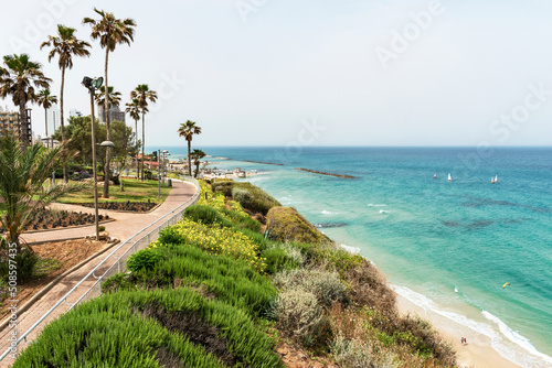 the promenade and resort hotels on a cliff high above the Mediterranean beach at Netanya in Israel with a couple walking, boats sailing and kitesurfing with a clear blue sky