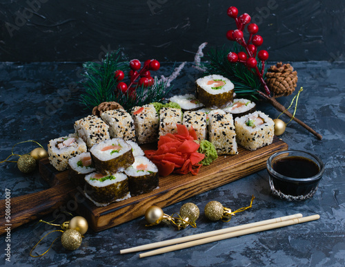 Large sushi set on a wooden board. Rolls with sesame seeds, nori, cheese, red fish, cucumber and eel, ginger, wasabi, soy sauce and christmas decor on a dark background