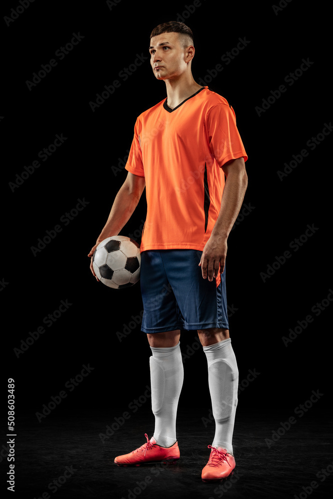 Young male soccer player in orange-blue football kit posing with ball isolated on dark background. Concept of sport, goals, competition, male hobby, occupations
